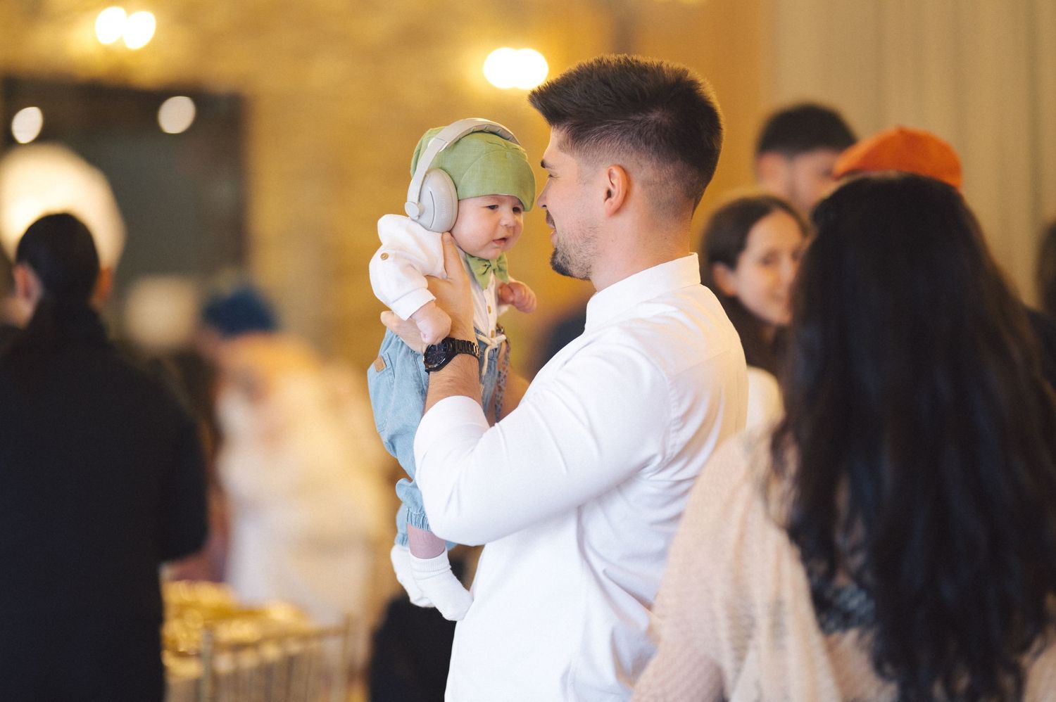 An adult in a white shirt holds a young child wearing a light blue outfit in an indoor event setting with warm lighting.