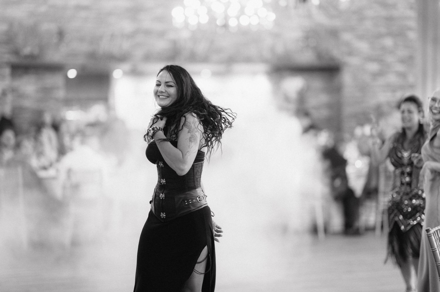 A graceful dancer with flowing hair performs in black and white at an indoor event with blurred figures in the background.