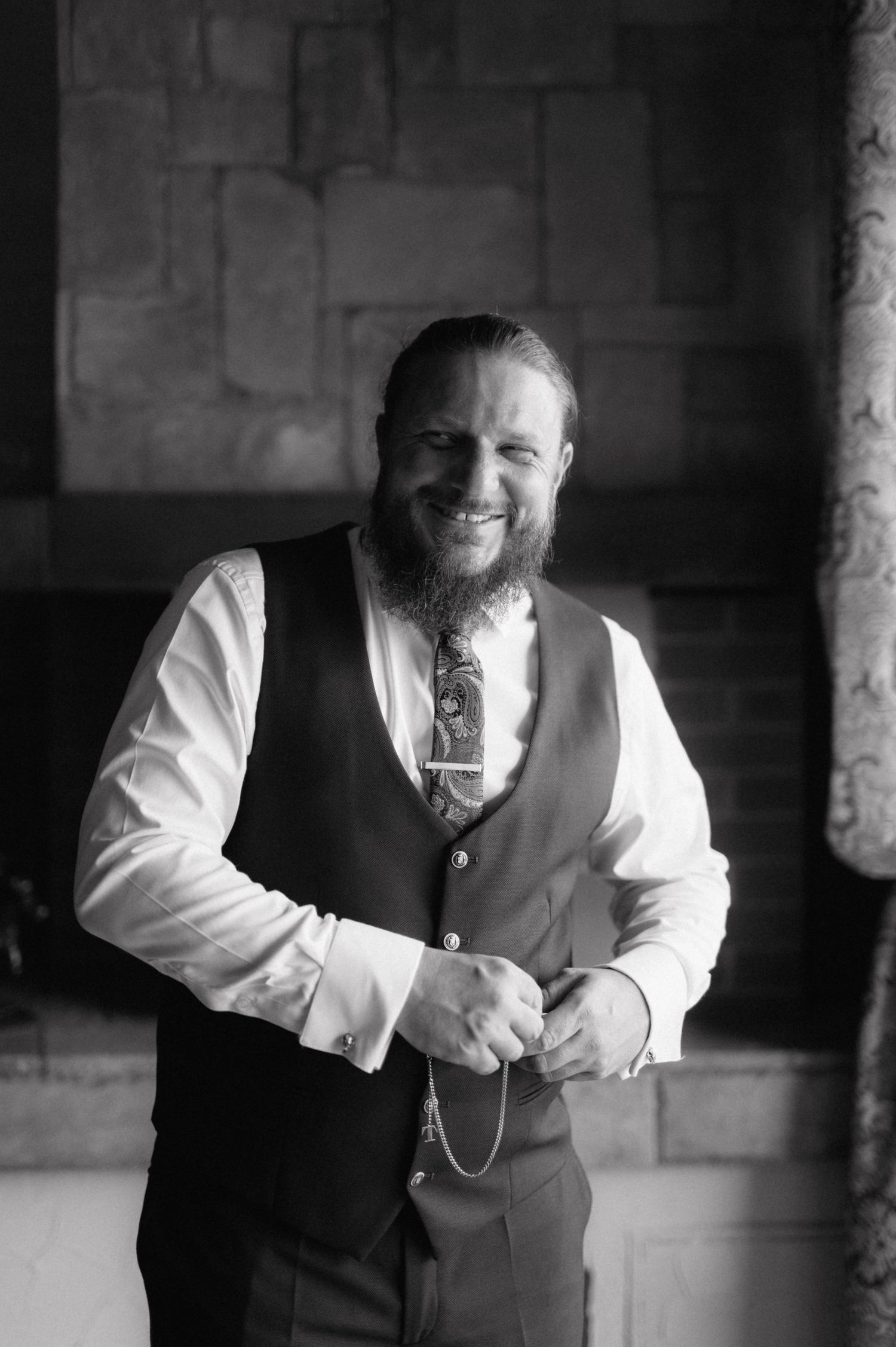 Black and white portrait of a bearded person wearing a dark vest and white dress shirt standing by a stone wall.