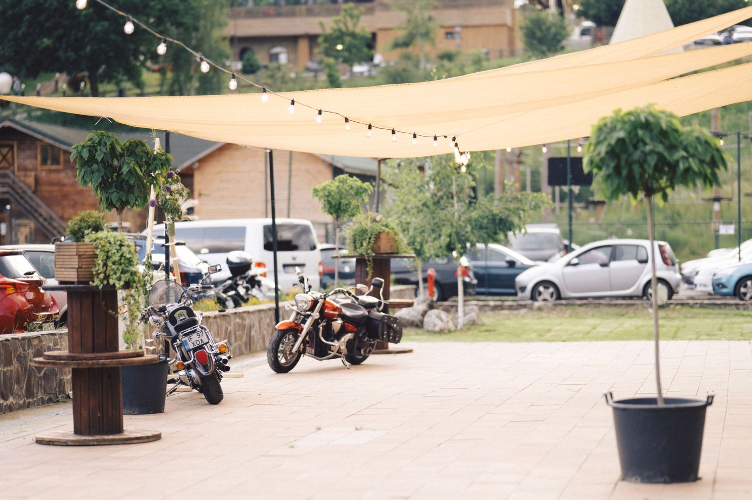 Motorcycles parked under a beige shade sail in an outdoor parking area with string lights and plants.