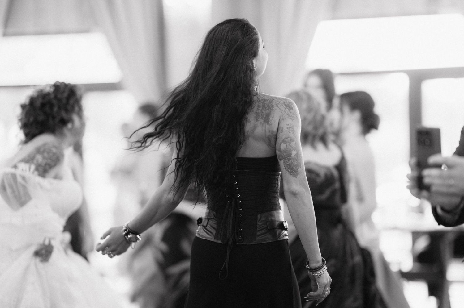 Black and white photo of person with long dark hair dancing at an indoor party or event, wearing a dark corset top.