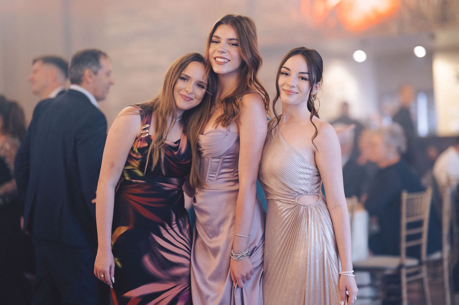 Three women in elegant evening gowns pose together at a formal event with soft lighting and blurred background decorations.