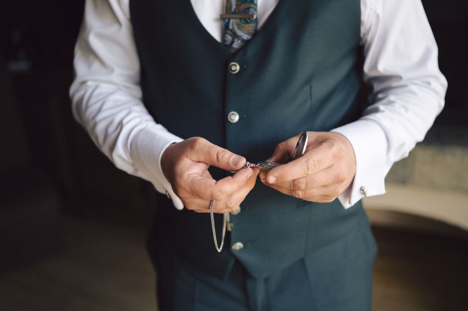 Close-up shot of hands holding a pocket watch while wearing a dark green vest and white dress shirt.