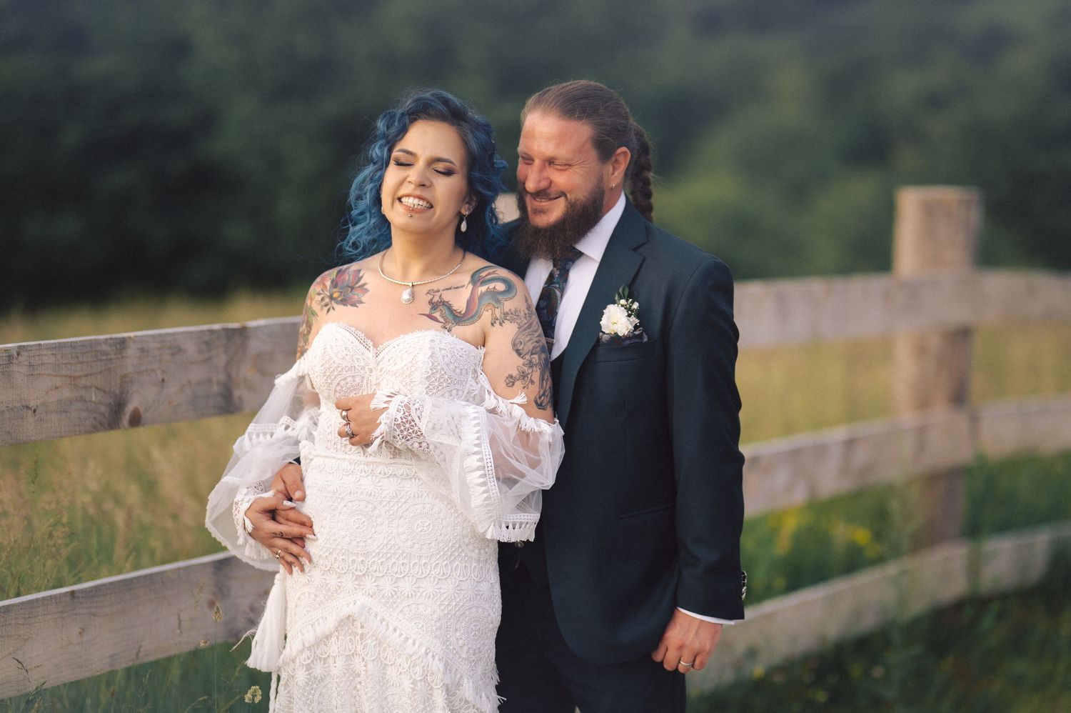 A newlywed couple shares a romantic moment together by a rustic wooden fence in a countryside setting.