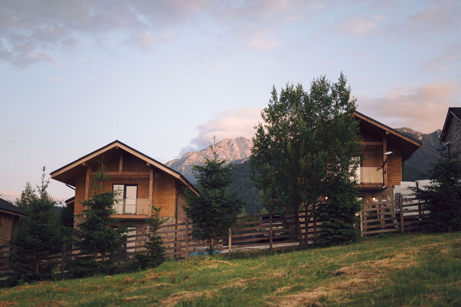 Rustic wooden cabins perched on a grassy hillside with mountain views and pink sunset clouds in the background.