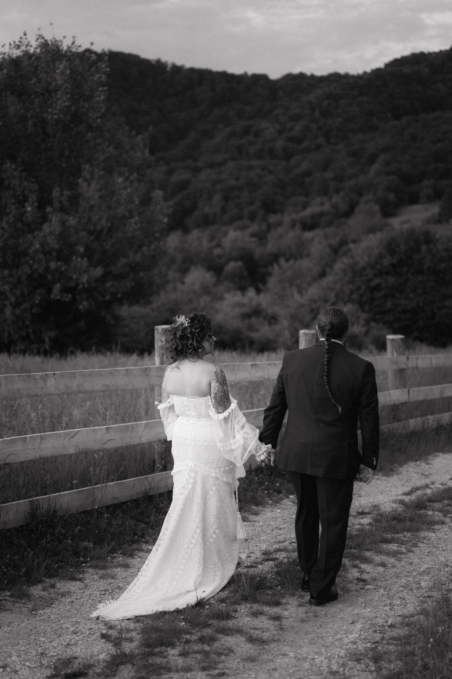 A black and white wedding photo series shows a couple walking together along a rustic fence-lined path.