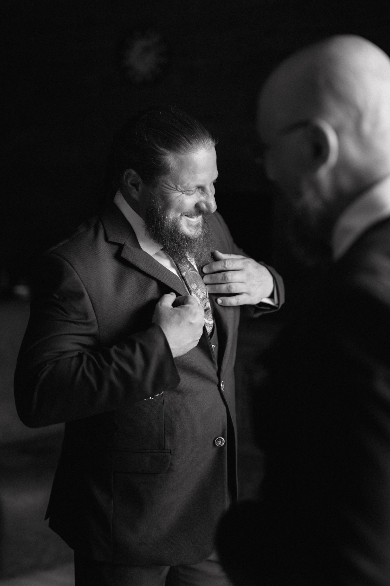 Black and white candid photo shows a suited man laughing joyfully while adjusting his tie during a formal event.
