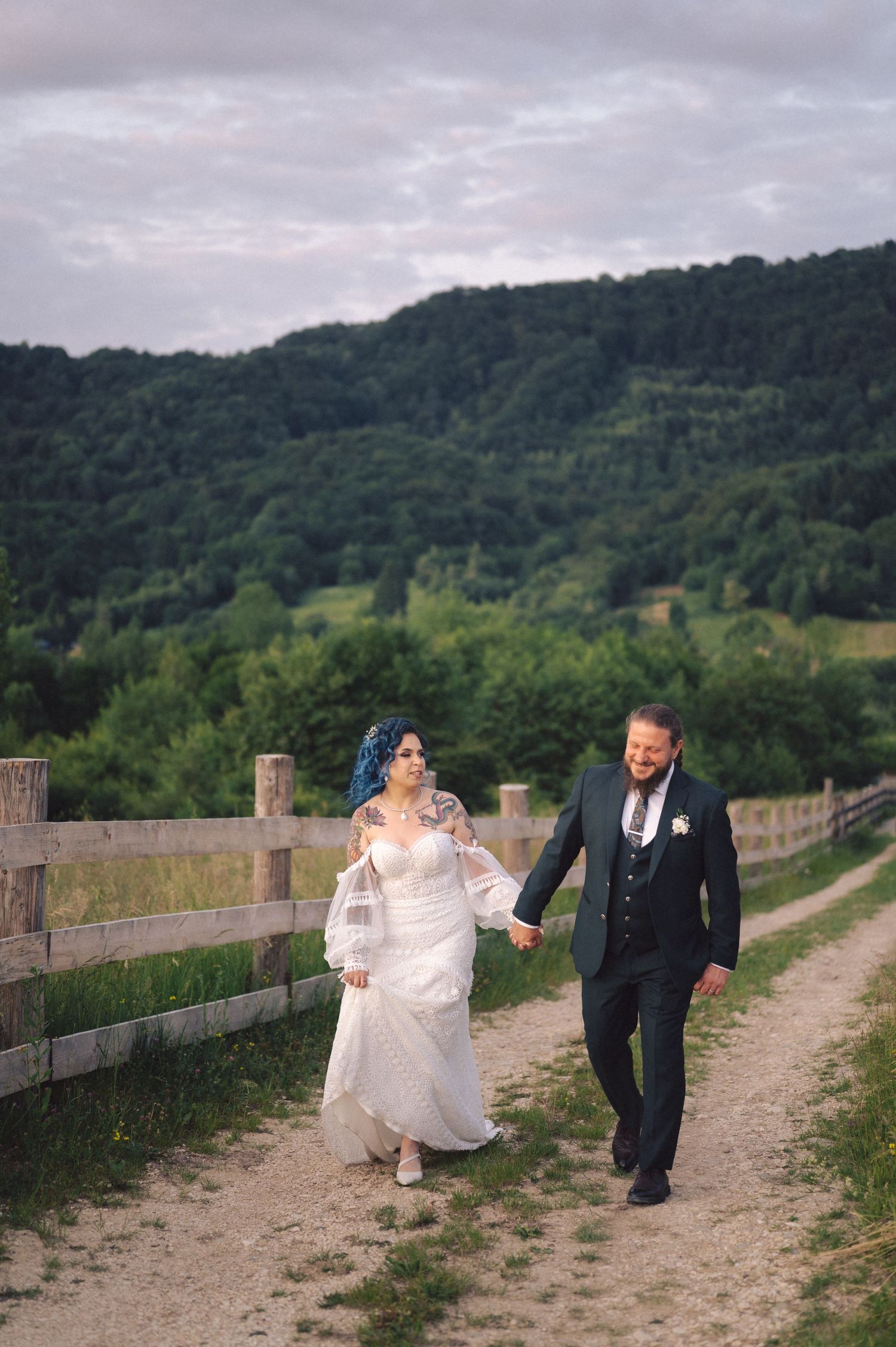 A newlywed couple walks hand in hand along a dirt path bordered by wooden fencing with rolling green hills in the background.