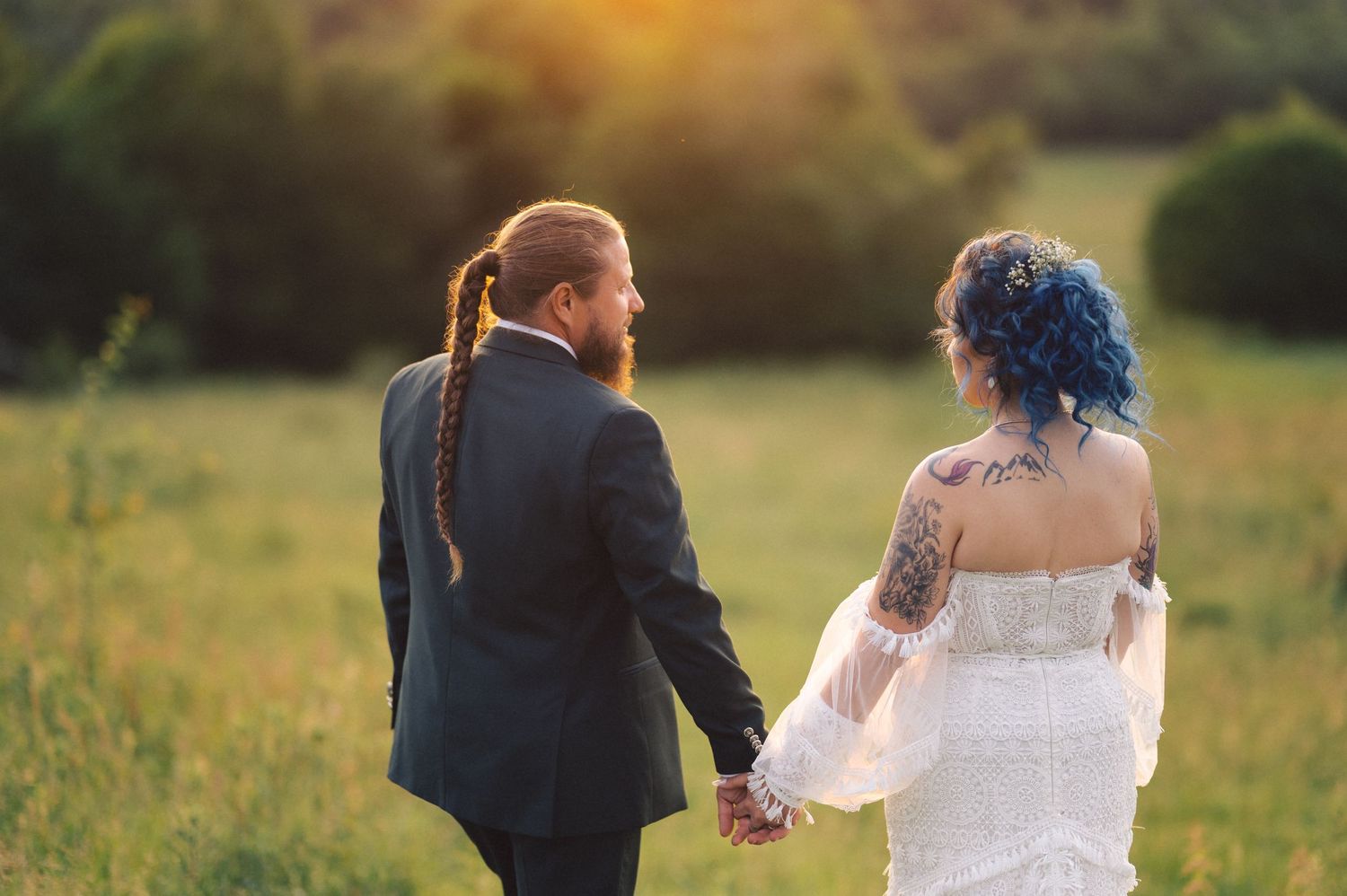A couple holds hands in a golden field during sunset while wearing formal wedding attire.