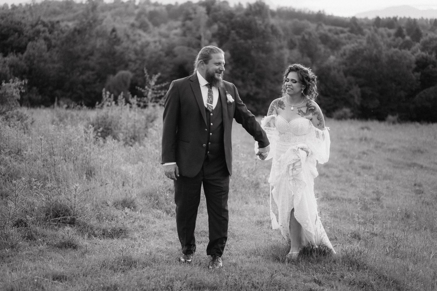 A couple in formal black and white attire walking together in a grassy meadow on a cloudy day.