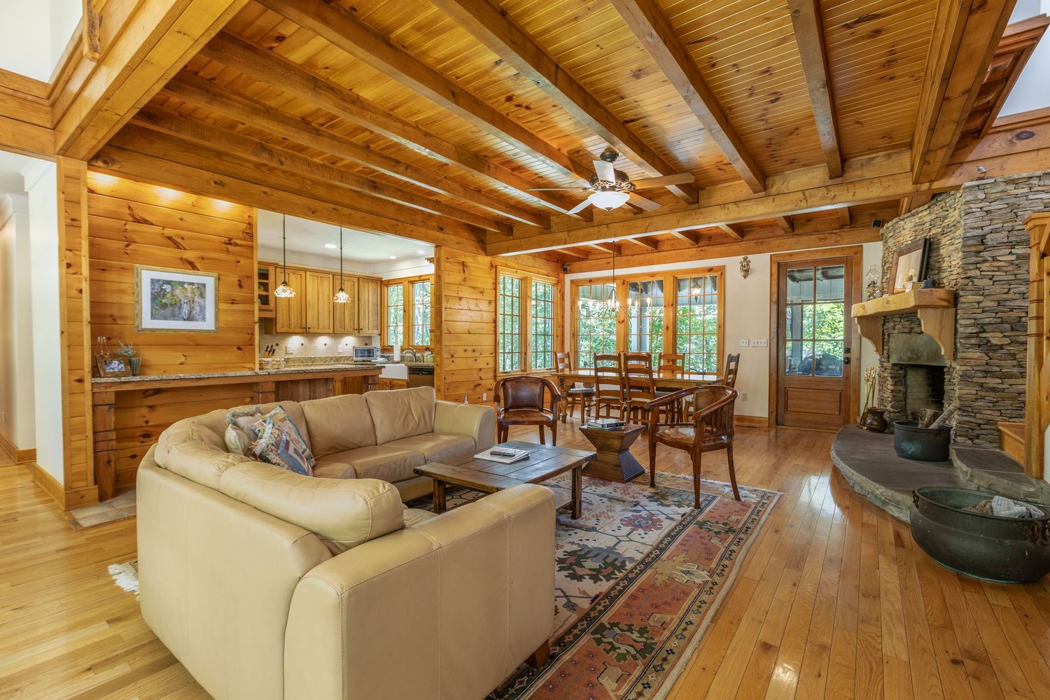 Luxurious log cabin living room with exposed wood beams, stone fireplace, and large windows showcasing forest views.