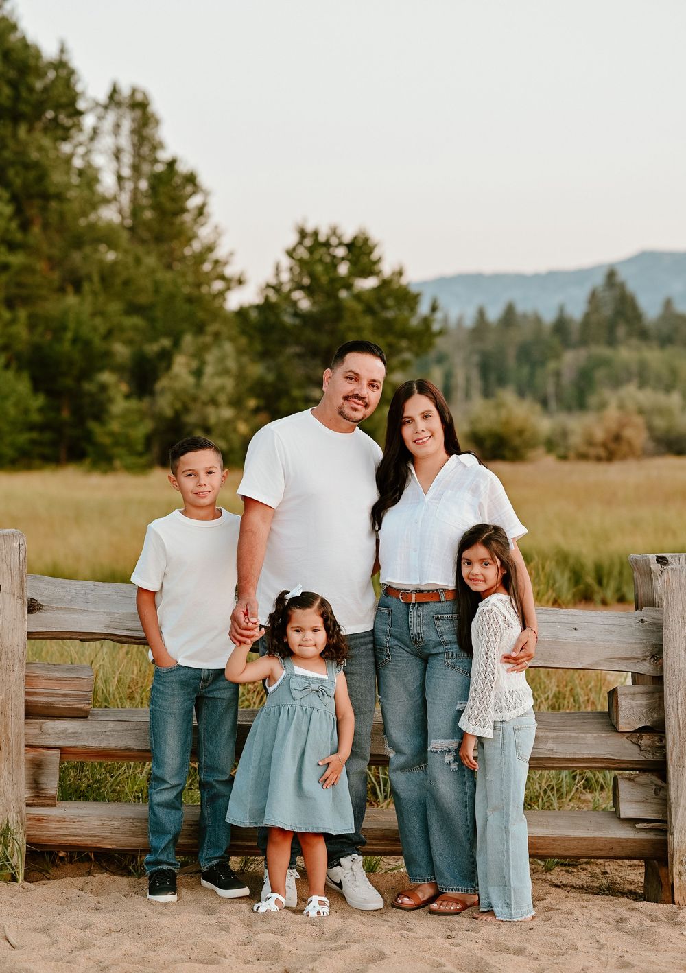 Family in coordinating white and blue outfits poses together on wooden fence in outdoor rustic setting at sunset.