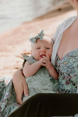 Baby wearing a mint green outfit with matching bow rests against someone in a floral dress outdoors.