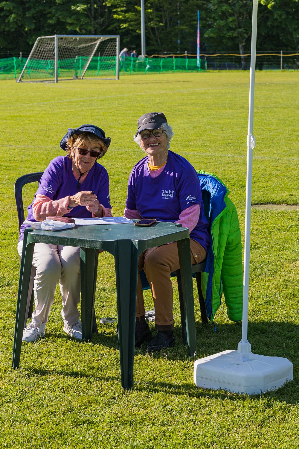 People seated at tables on a sunny day watching an outdoor event on a green sports field.