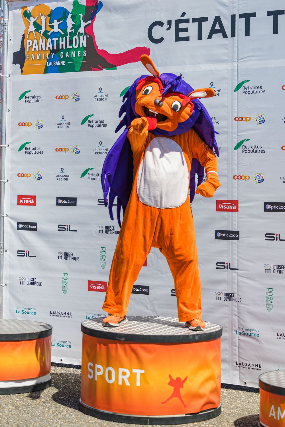 Orange and purple mascot character poses on a sports podium at Panathlon event with sponsor backdrop.