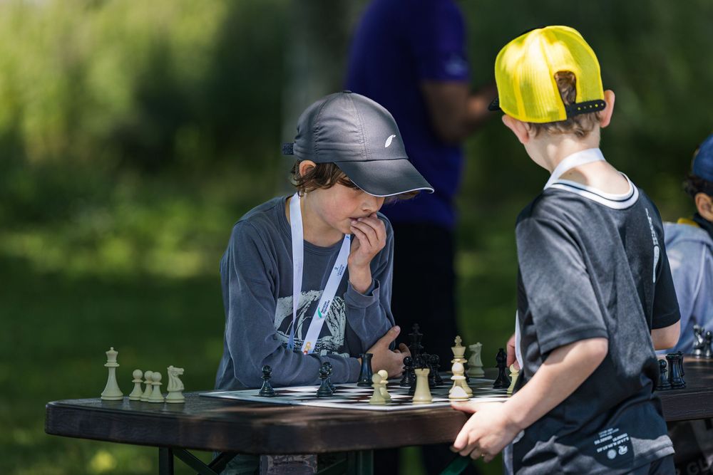 Two people playing chess outdoors at a table wearing baseball caps and grey hoodies.