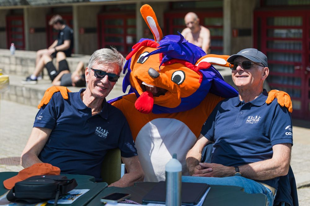 Two staff members in navy blue uniforms pose with an orange and blue team mascot at a sporting event.