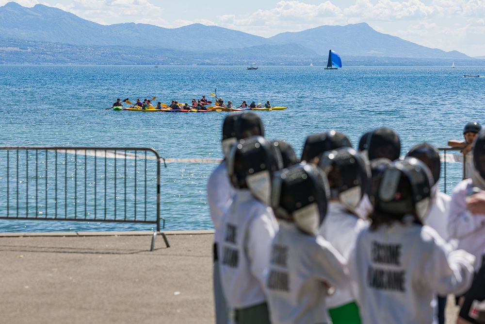 People in white uniforms watch kayakers paddling in blue waters with mountains and a sailboat in the distance.