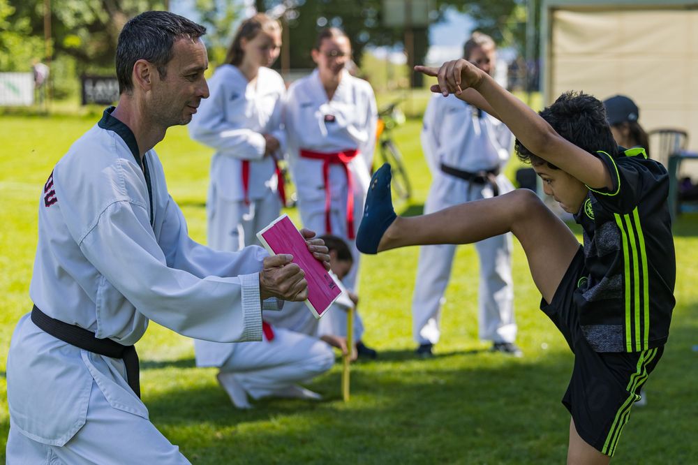 Two martial arts practitioners demonstrate a high kick during an outdoor training session in a grassy area.