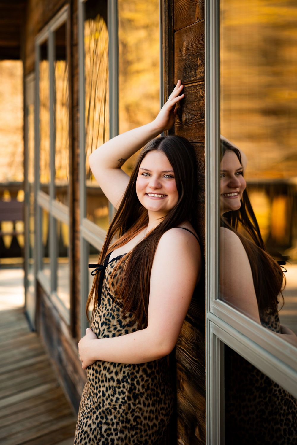 Reflection in rustic wooden window shows someone in leopard print dress posing against warm golden lighting.