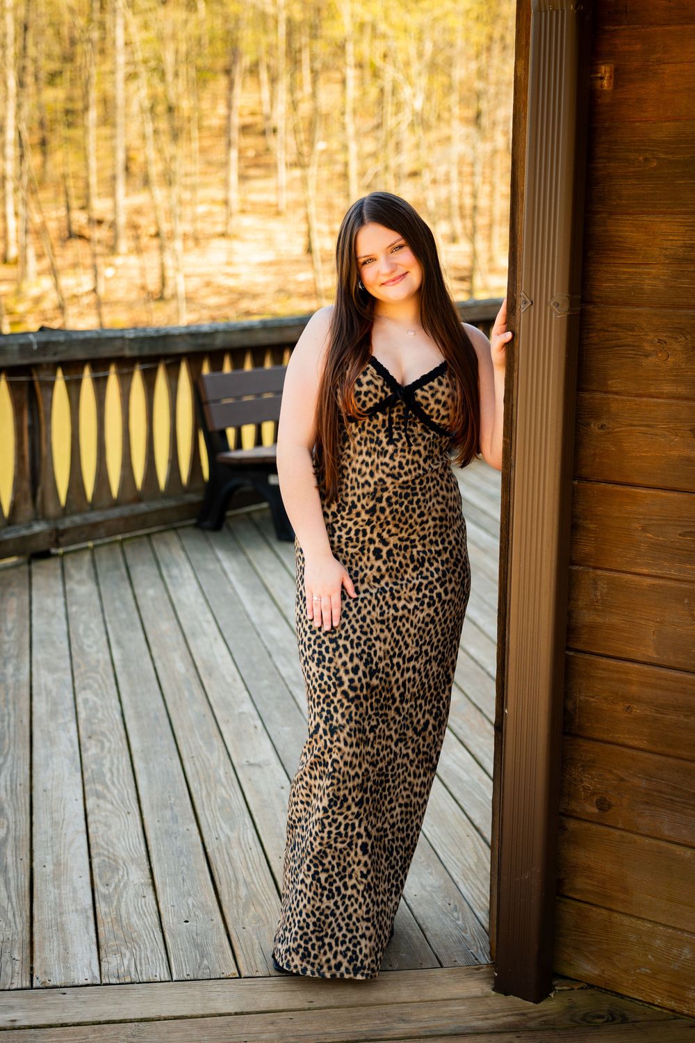 A person in a leopard print maxi dress poses on a wooden deck surrounded by autumn woods.