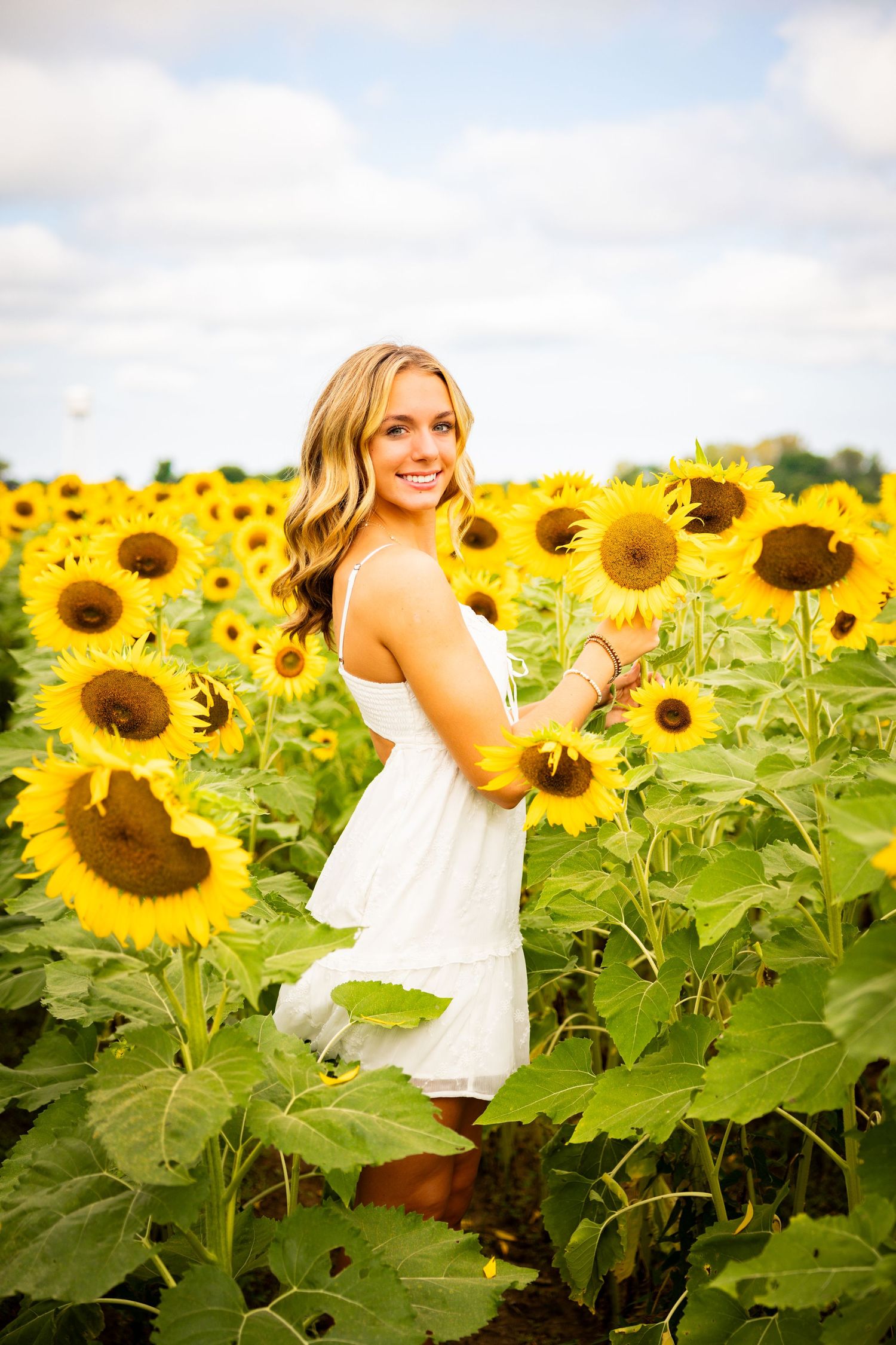 A person wearing a white dress stands in a vibrant field of bright yellow sunflowers on a sunny summer day.