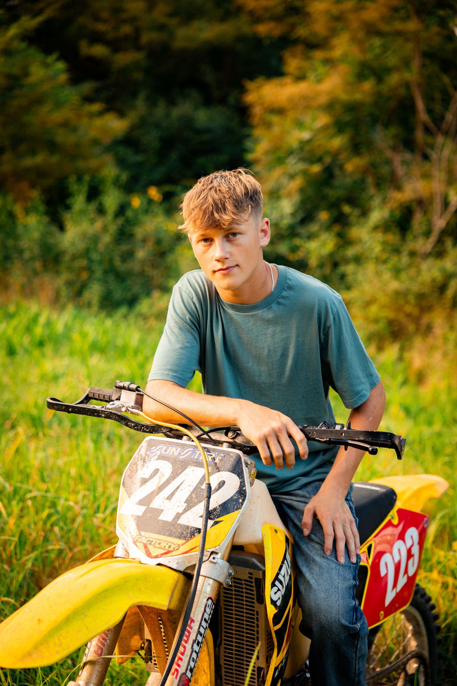 A person sits on a yellow dirt bike outdoors during golden hour with autumn foliage in the background.