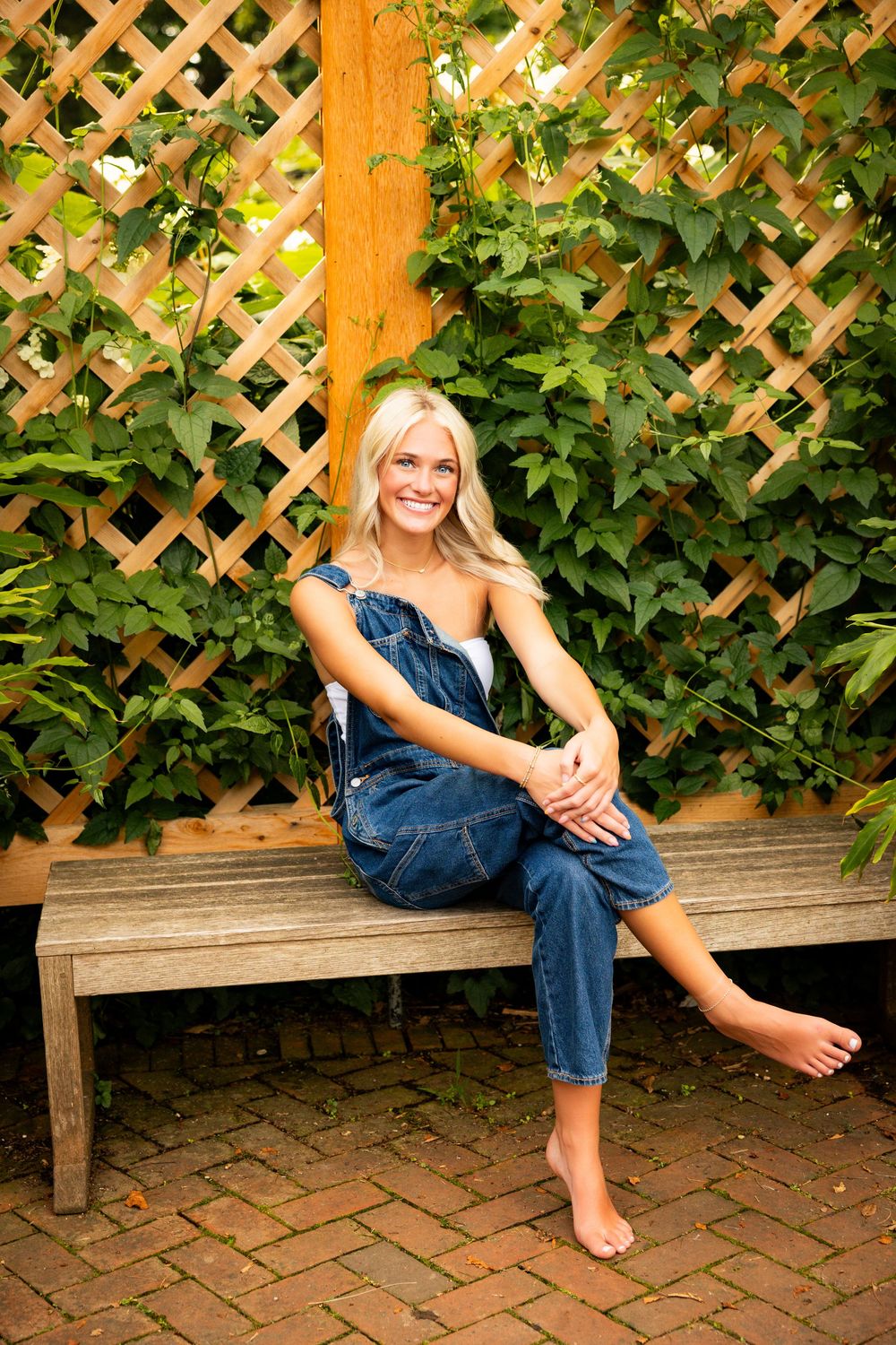 Someone in denim overalls sits barefoot on a wooden bench against a lattice fence with climbing vines and flowers.