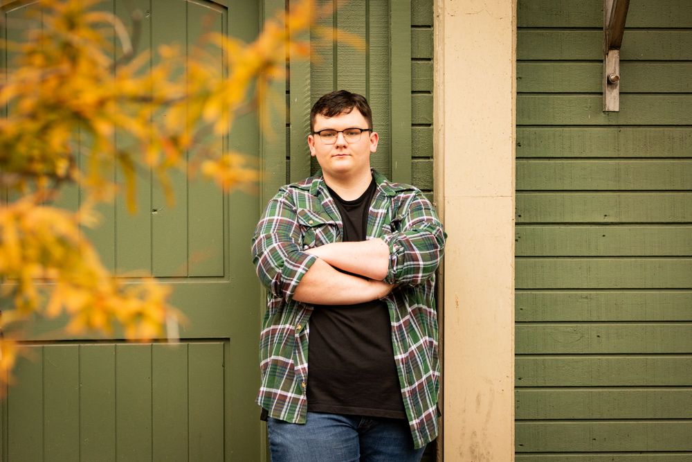 A person in a plaid flannel shirt and jeans stands with arms crossed against a green door with autumn foliage in view.