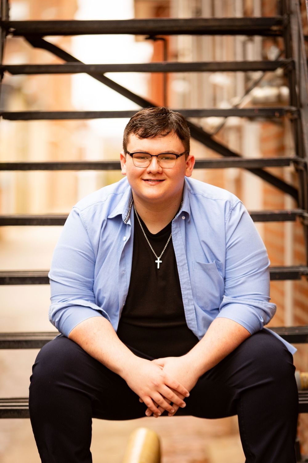 A person sits on outdoor stairs wearing a light blue button-up shirt over a black v-neck with a cross necklace.