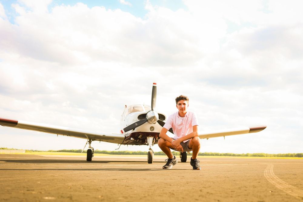 A person in casual clothing crouches on a sunny tarmac next to a small private airplane.