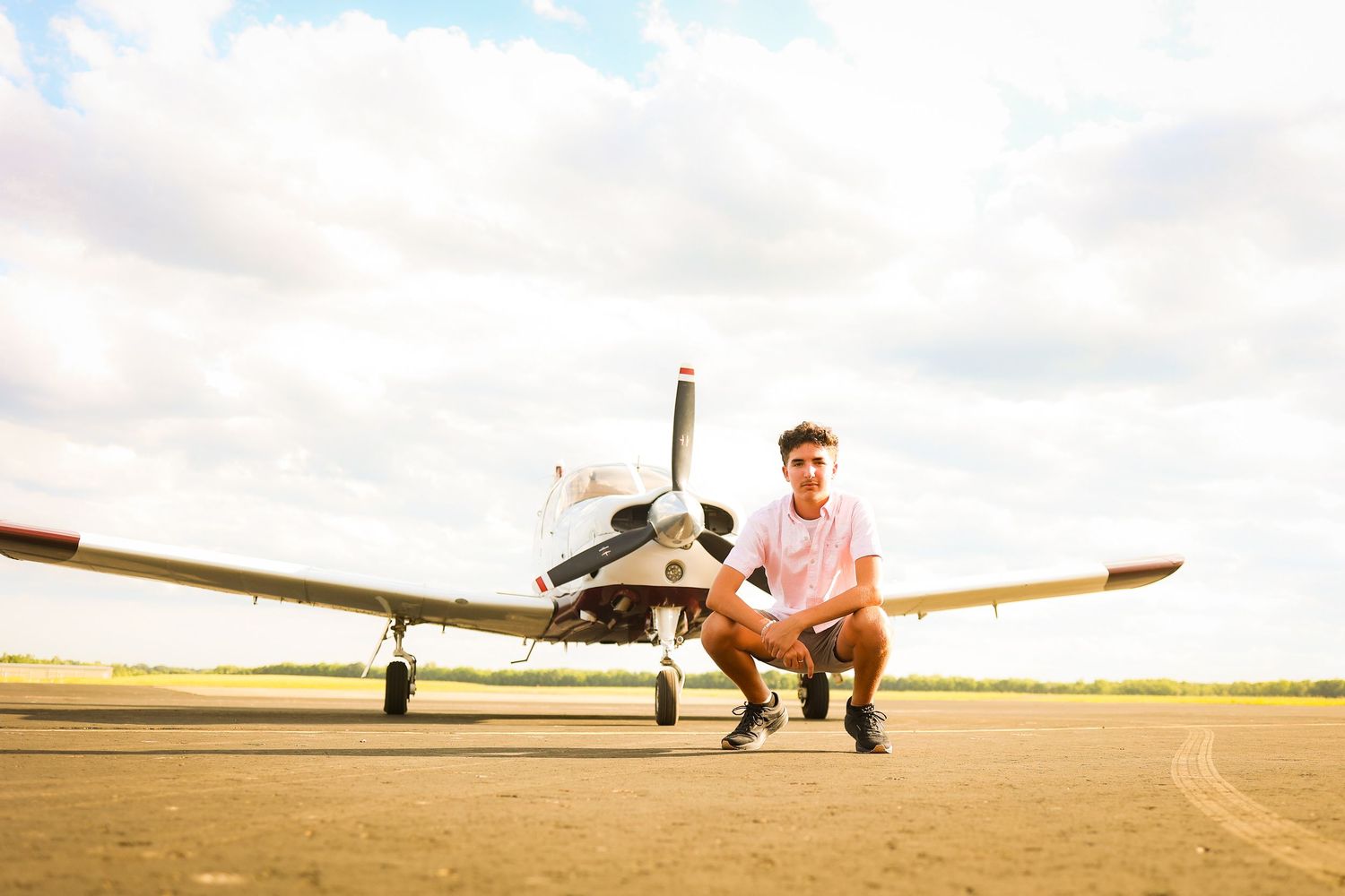A person in casual clothing crouches on a sunny tarmac next to a small private airplane.