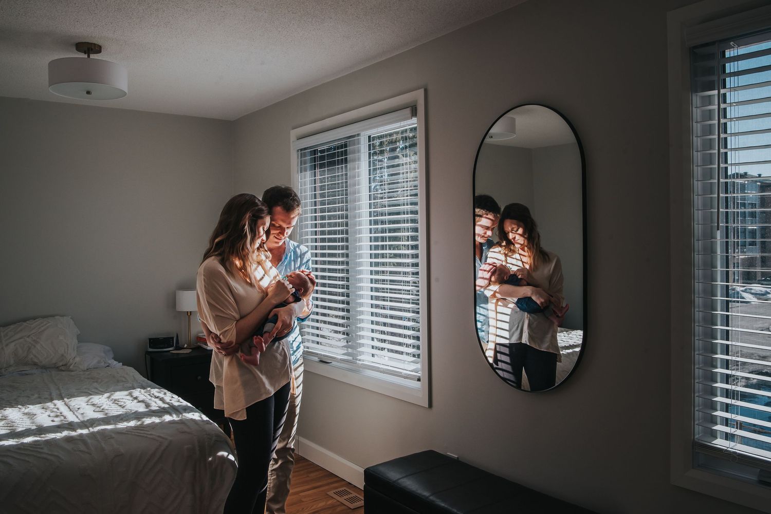 Parents and baby stand by a window while their reflection appears in a wall mirror in a sunlit bedroom.