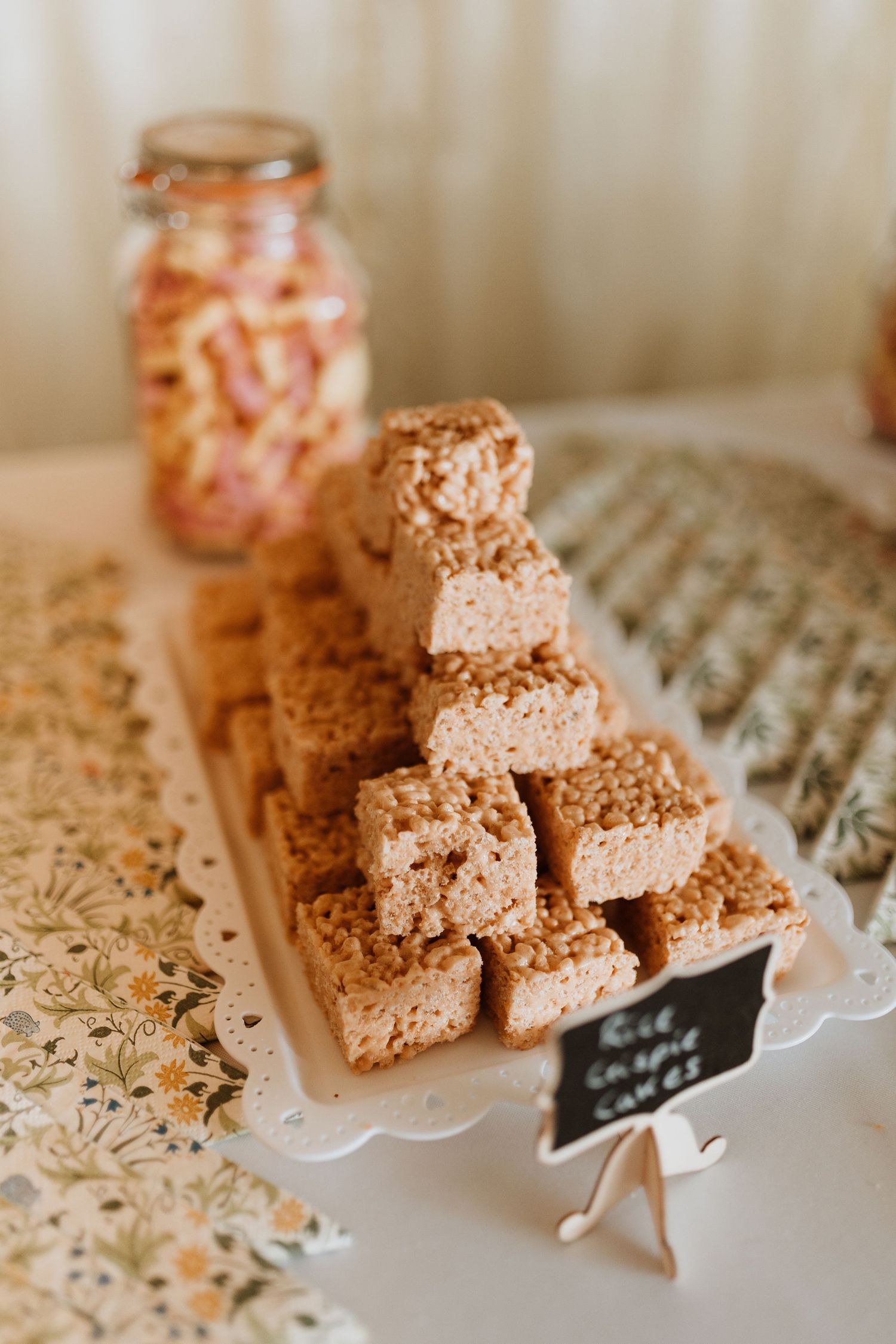 Stacked rice krispie treats dusted with cocoa powder displayed on a wooden board next to a small jar of preserves.