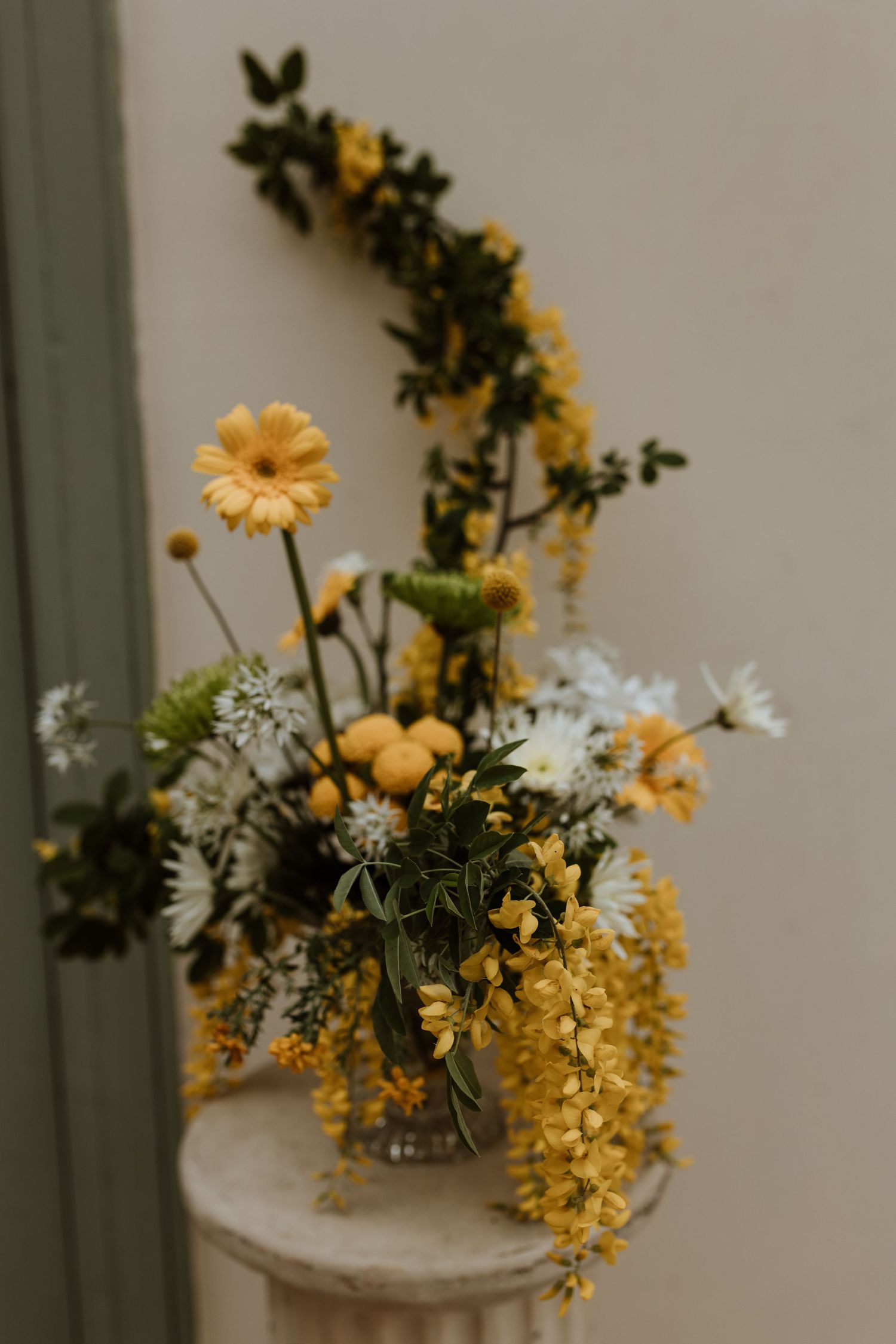 A moody floral arrangement with yellow daisies and white flowers in a vase on a light colored pedestal.