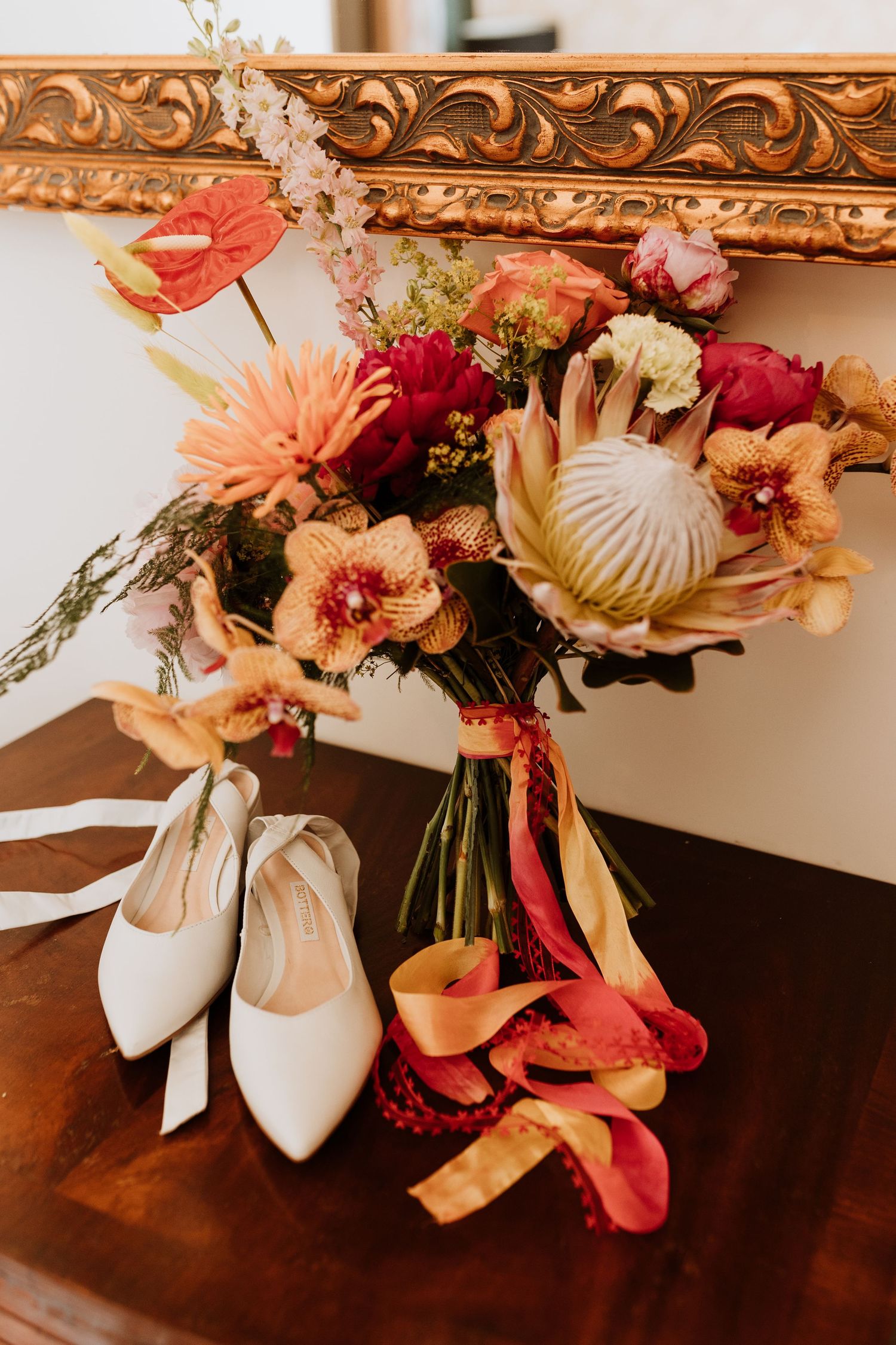 White bridal shoes with satin bows displayed with coral and burgundy floral arrangements on a dark wooden surface.