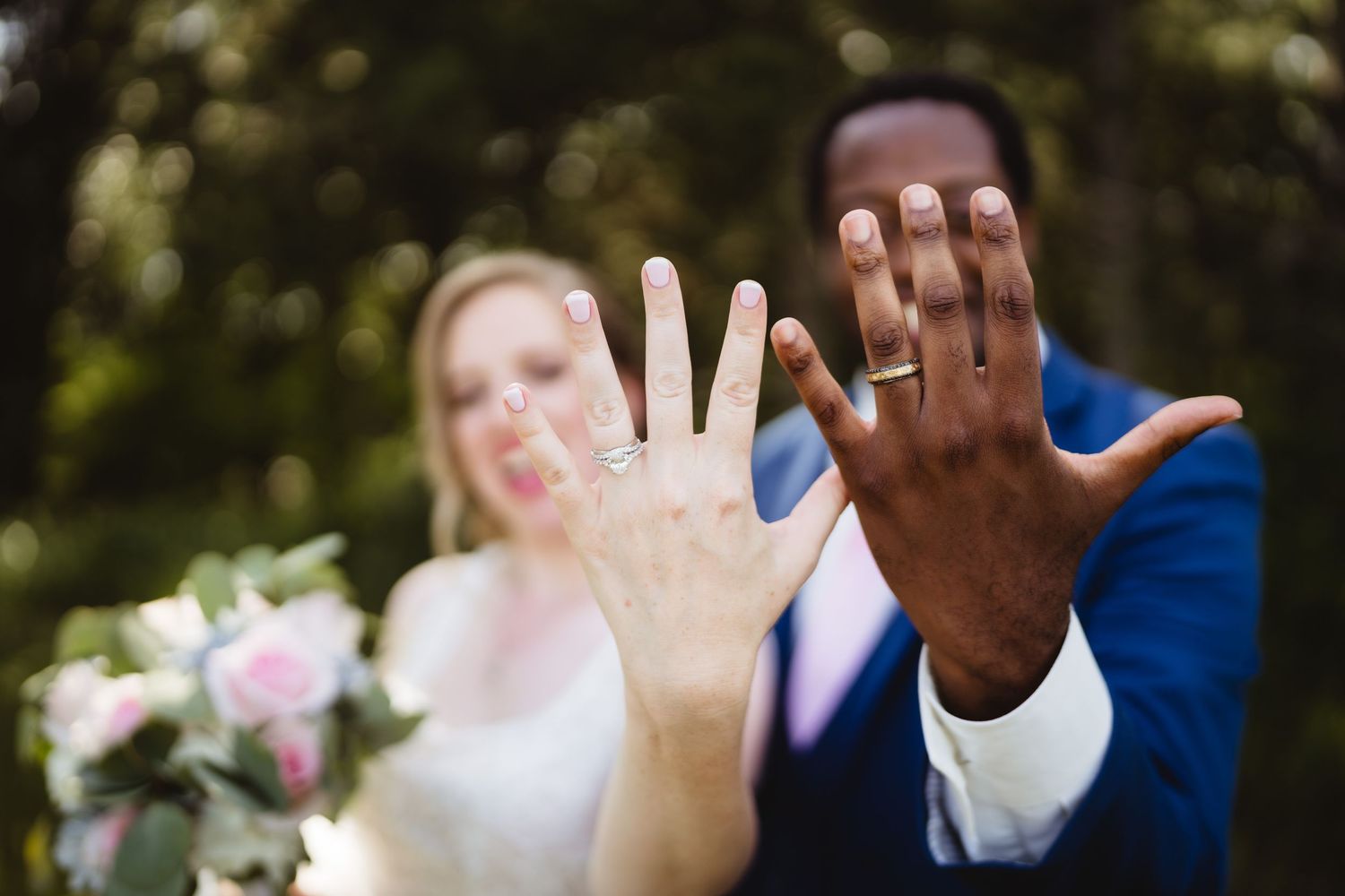 Two hands displaying wedding rings against a blurred natural background with pink flowers.