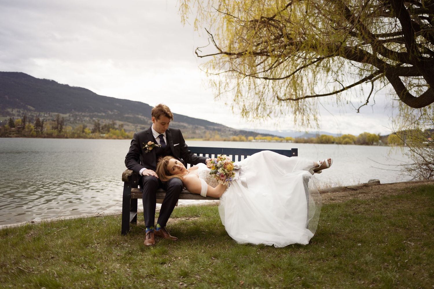 Playful wedding portrait by a lakeside with couple on rustic bench.