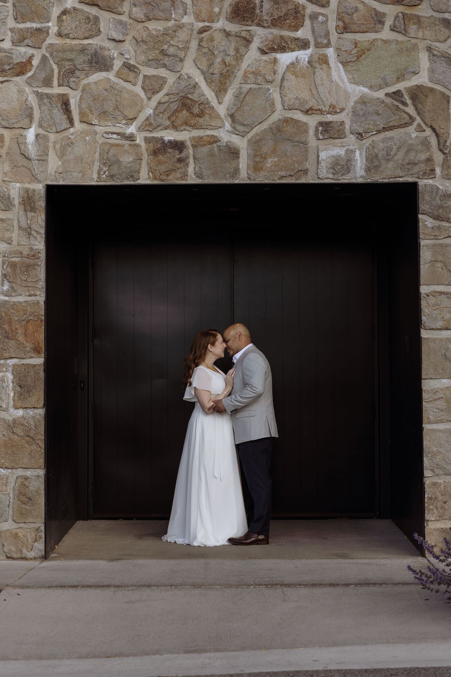 Newlyweds share intimate moment in stone archway with dark wooden door.