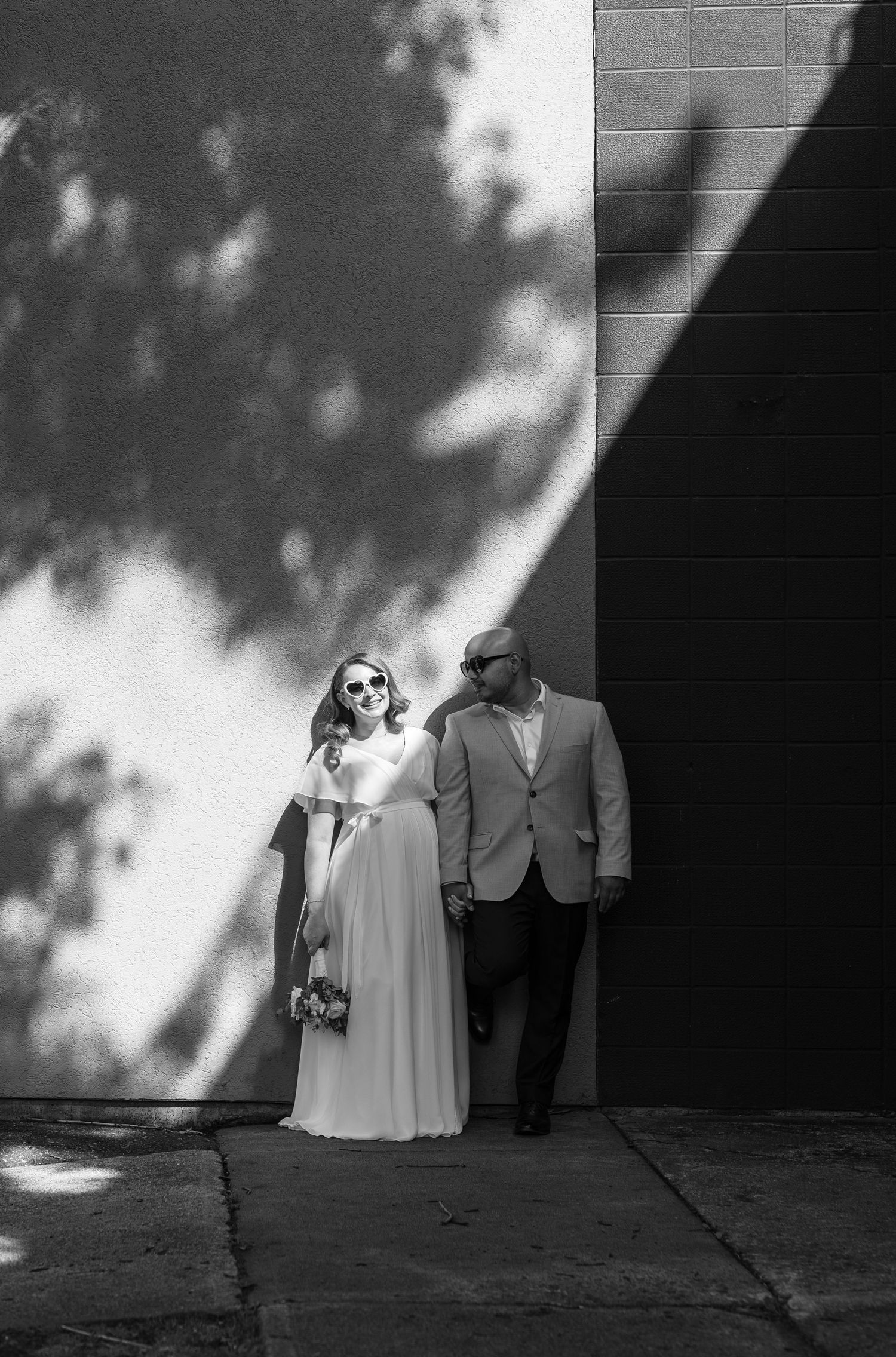 Black and white wedding photo with artistic tree shadow patterns on a wall as a couple stands together.