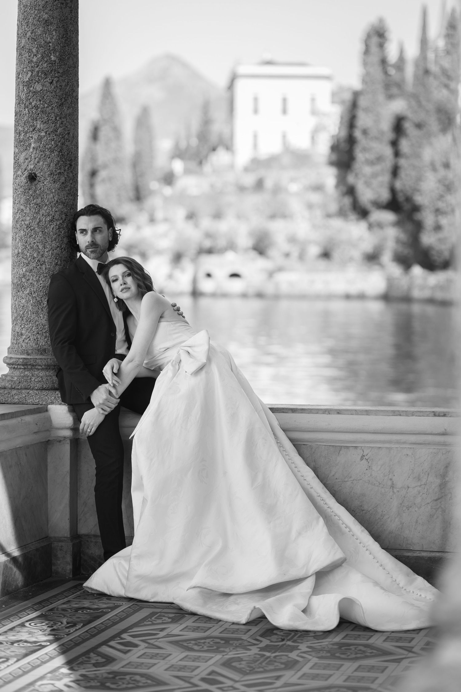 Elegant wedding portrait by lakeside with classic architecture and mountain backdrop.