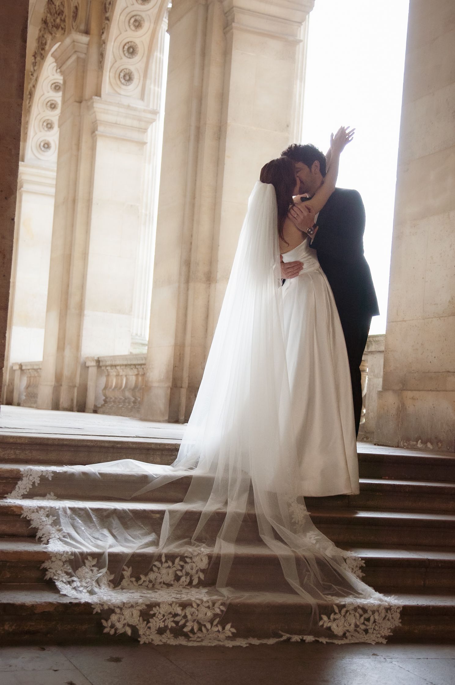 Wedding couple shares intimate moment on grand stone staircase with flowing lace veil.