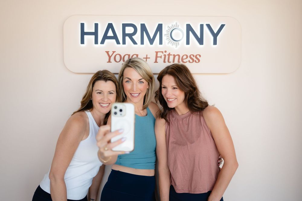Three people take a selfie together while standing in front of a Harmony Yoga and Fitness studio sign.