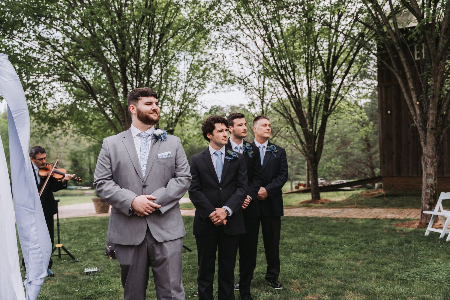 A wedding party poses outdoors in a garden setting with groomsmen in gray and black suits and bridesmaids in light dresses.