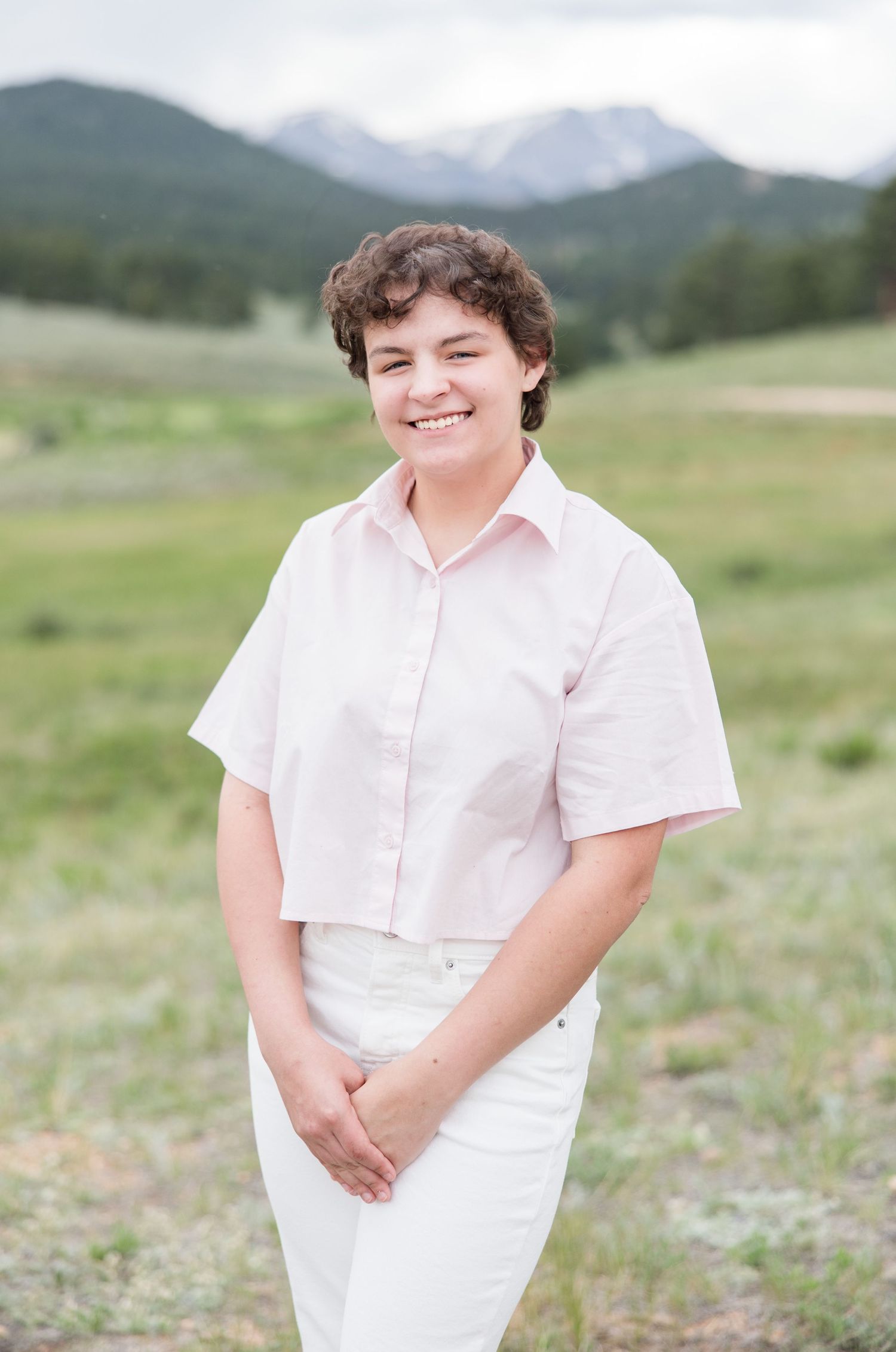 Someone in a light pink button-down shirt and white pants smiles while standing in a mountain meadow landscape.