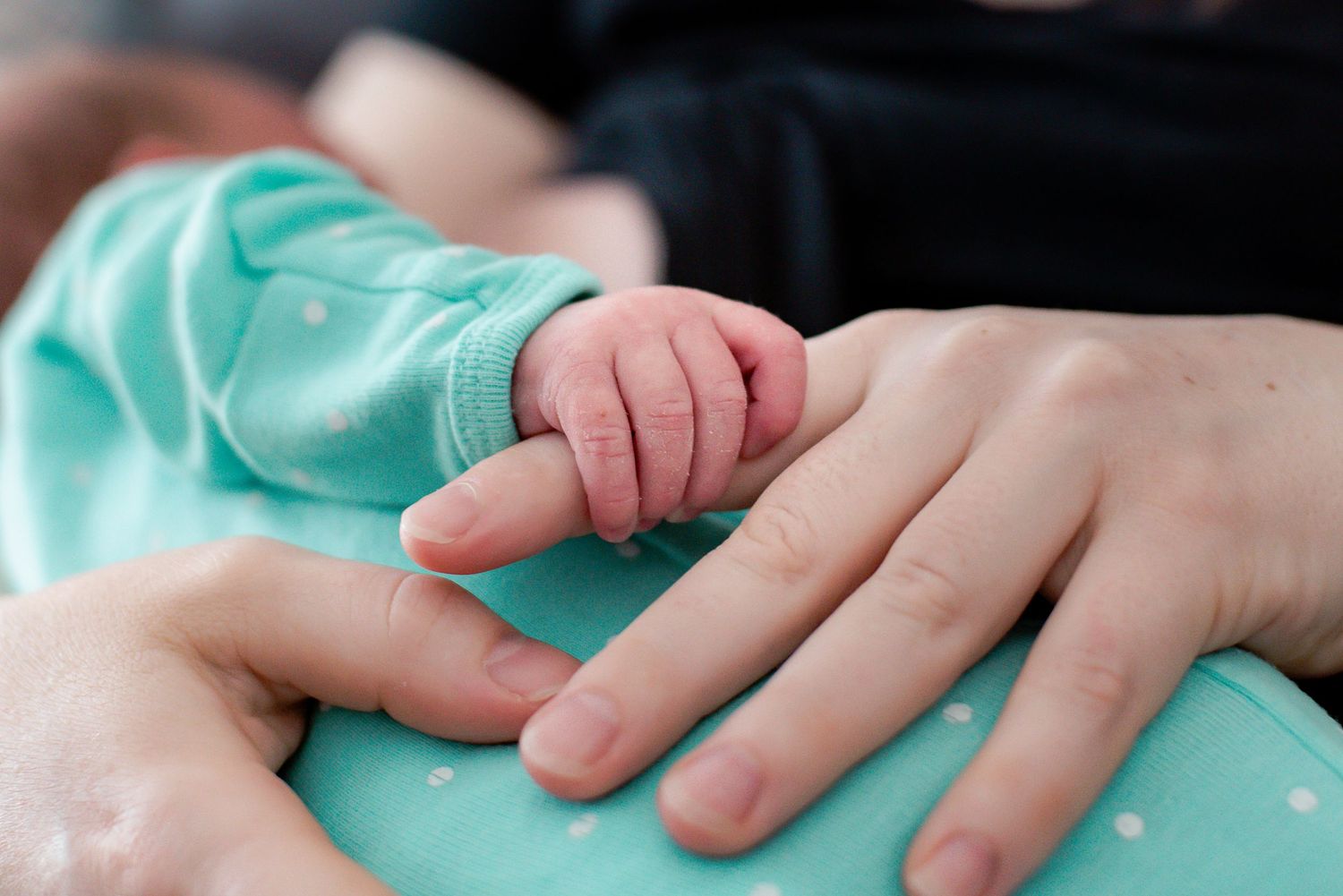 A tiny baby hand gently grasping an adult finger against a turquoise fabric background.