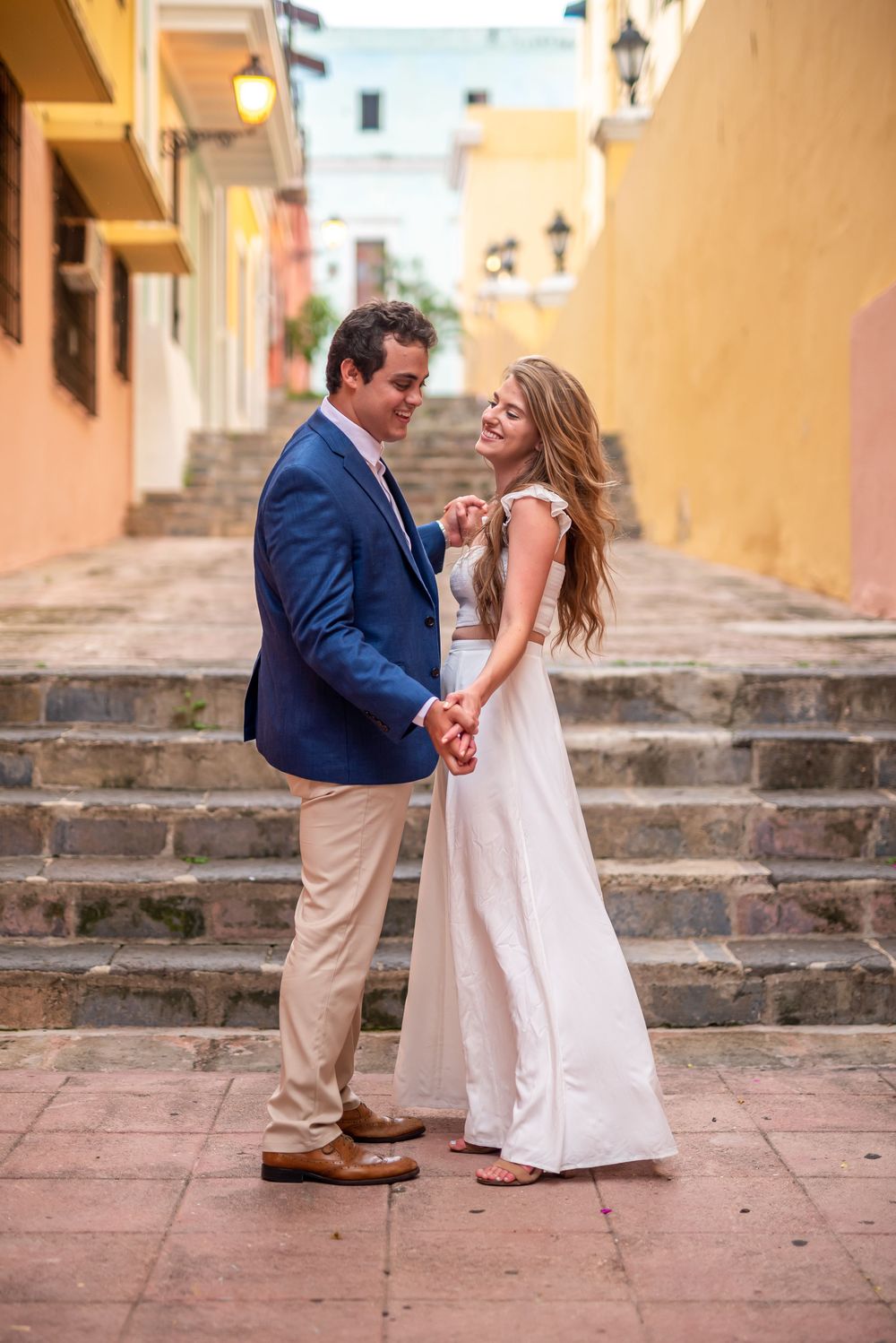 A couple shares a romantic moment on stone steps in a charming alleyway with yellow walls and soft lighting.