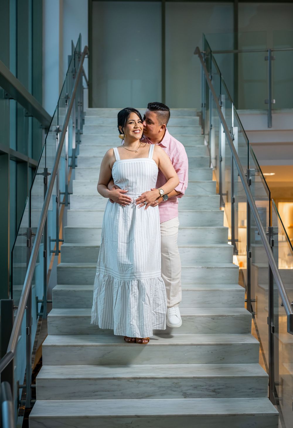 A couple shares an intimate moment on a modern staircase, with her in a white dress and him in pink.