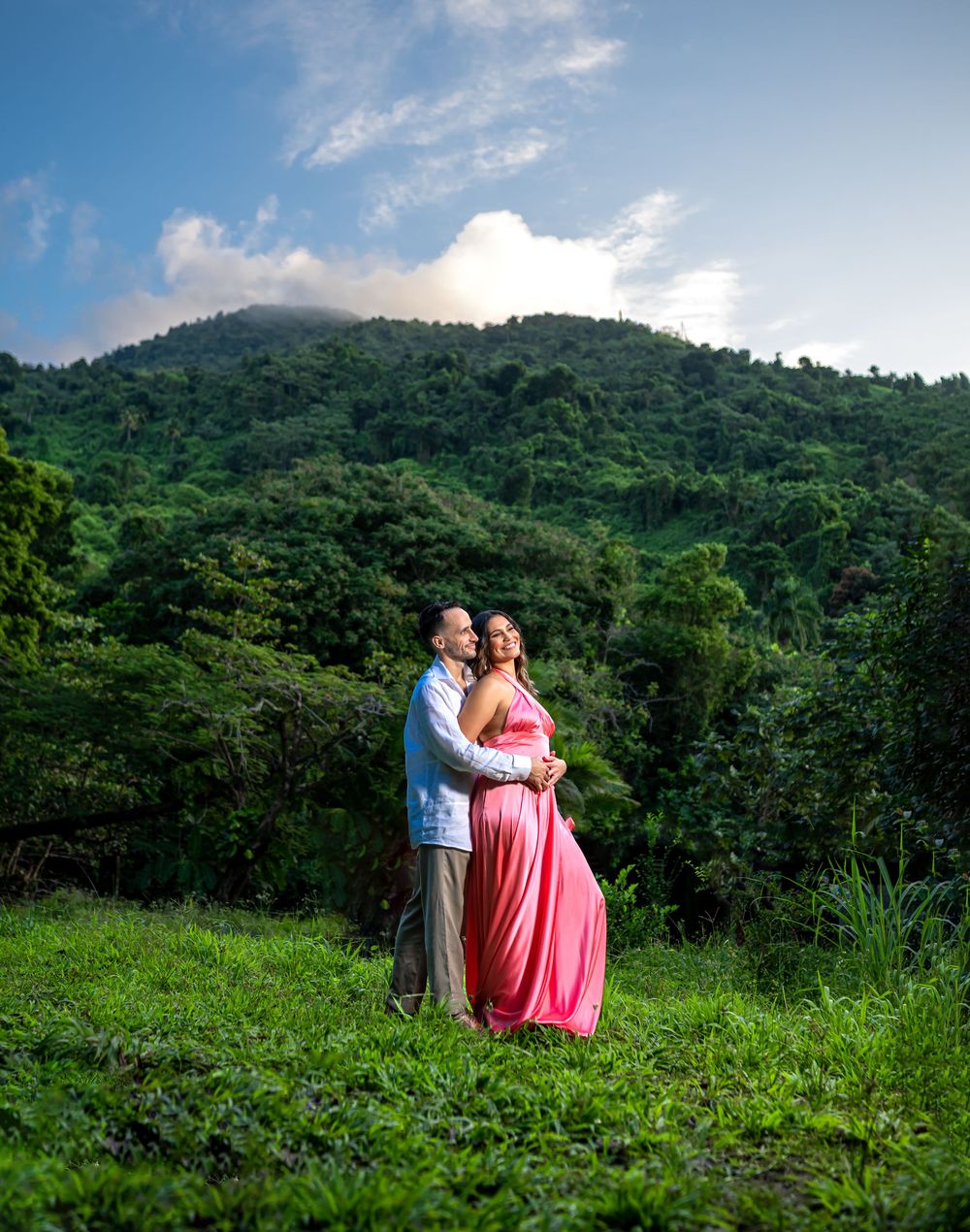 A romantic couple poses together on a lush green hillside with mountains and tropical forest in the background.