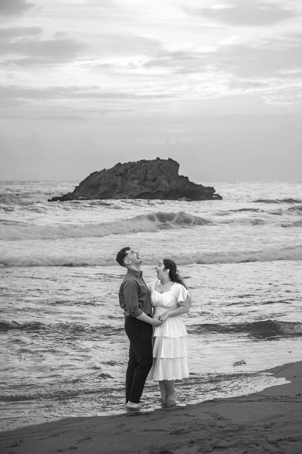 A couple shares a romantic moment on a misty beach with a dramatic rocky outcrop in the ocean behind them.
